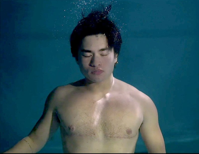 An Asian person floats upright underwater, his eyes closed and hair flowing in the water. He's shirtless and has top surgery scars across his chest.