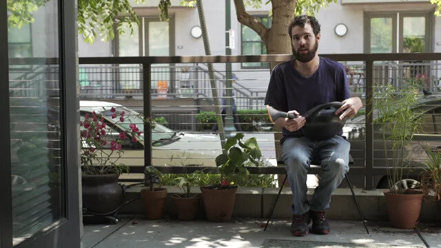 A man sits on a patio surrounded by plants. He has light skin, brown hair and a beard. He is wearing a dark t-shirt and jeans. He is holding a watering can.

